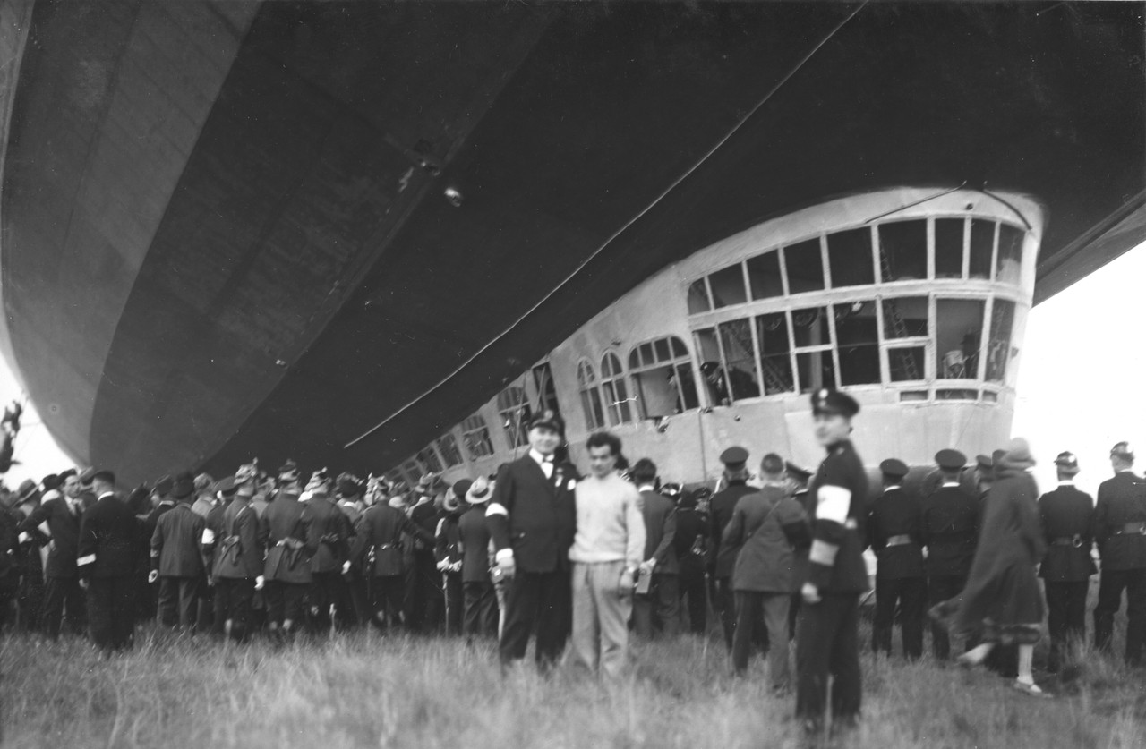 Um das Luftschiff und die Passagiere vor den Schaulustigen zu schützen, wurde der Zeppelin von Ordnungskräften abgeschirmt. Gut zu sehen ist hier die Führergondel mit der Kommandobrücke, in der sich außerdem eine moderne Funkstation, die zehn Kabinen der max. 24 Passagiere, ein Aufenthaltsraum, Toiletten und Waschräume sowie die Küche befanden. Dortmund, 10.8.1930 (Foto:  Hermann Tell, LWL-Volkskundearchiv)