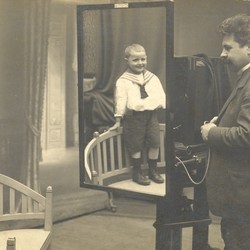 Wilhelm Tell steht auf einem Stuhl, ihm gegenüber ein Spiegel mit seinem Spiegelbild. Sein Vater steht an einer Fotokamera und fotografiert Wilhelm (vergrößerte Bildansicht wird geöffnet)