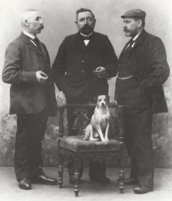 Schwarzweißfotografie von drei Männern im Anzug, die sich um einen auf einem Stuhl sitzenden Hund positioniert haben.