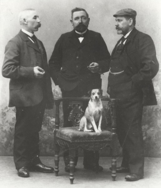 Schwarzweißfotografie von drei Männern im Anzug, die sich um einen auf einem Stuhl sitzenden Hund positioniert haben.