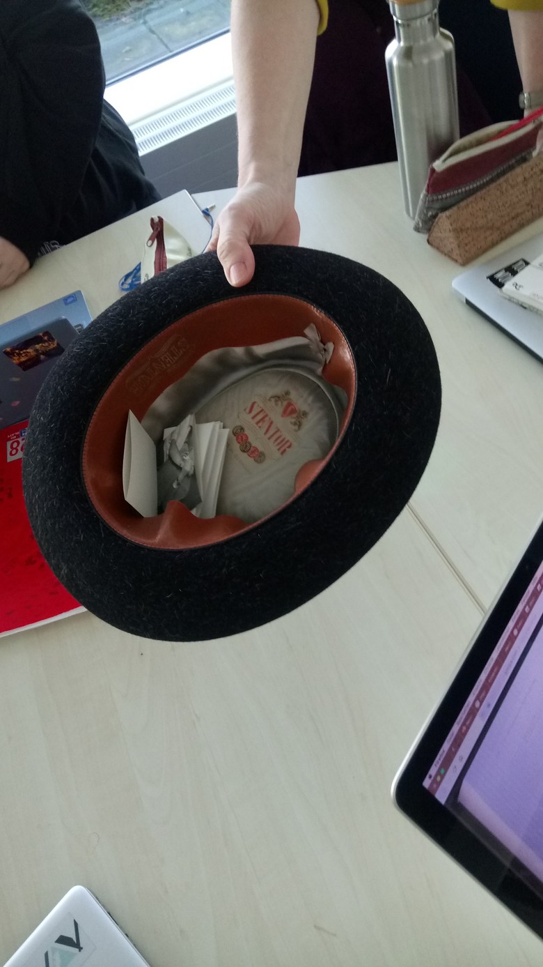 Ein Hut, in dem sich einige gefaltete Zettel befinden. Eine Hand hält den Hut über einem Tisch, auf dem sich Laptops und Schreibutensilien befinden.