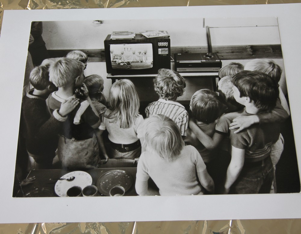 Ein Bild auf goldenem Hintergrund. Zu sehen sind mehrere Kinder vor einen Fernseher. Archiv für Alltagskultur.