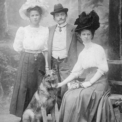 Zwei Frauen und ein Mann wurden gemeinsam mit einem Schäferhund vor einem waldähnlichen Hintergrund fotografiert. (vergrößerte Bildansicht wird geöffnet)