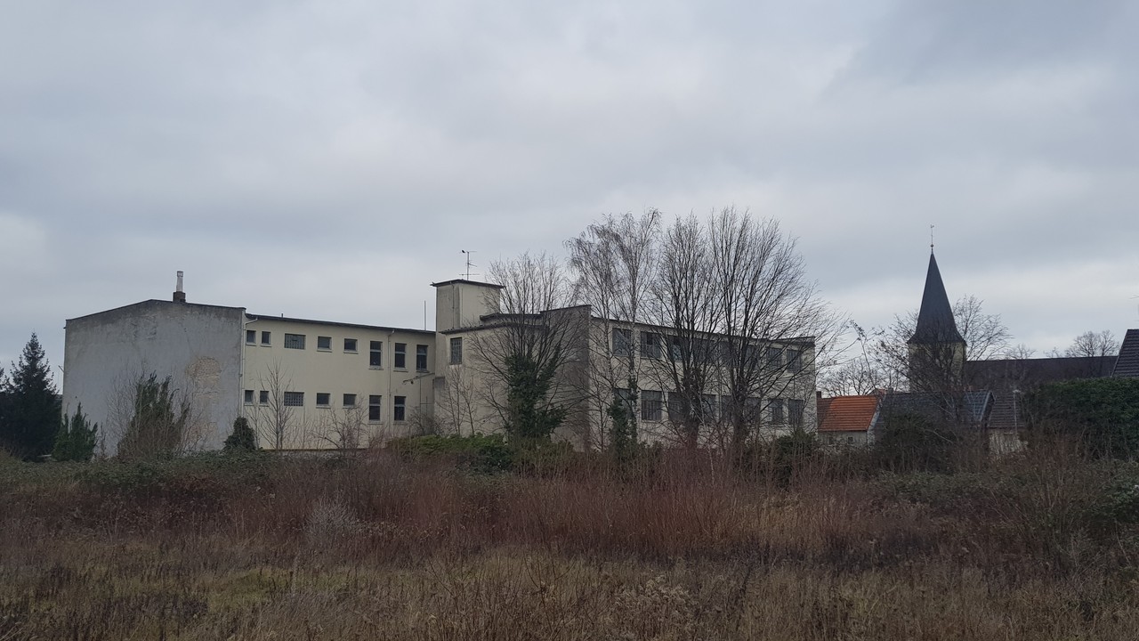 Das Gebäude, in dem das neue Depot in Lippstadt eingerichtet wird. Es handelt sich um ein großes Gebäude, dessen heller Putz abbröckelt. Im Vordergrund des Fotos befinden sich Bäume und Sträucher.