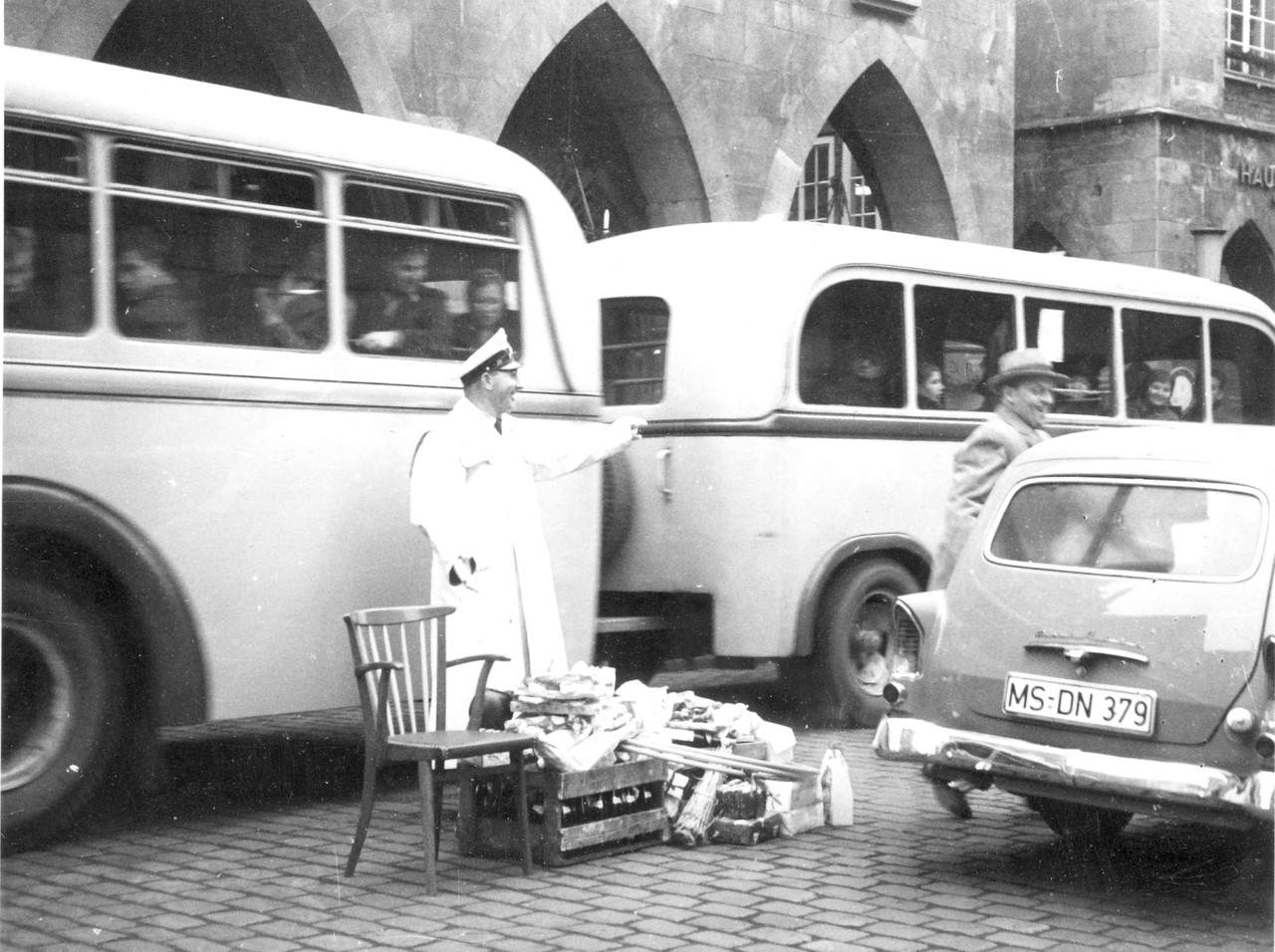 Im dichten Autoverkehr steht der Verkehrspolizist mit einem Berg an Geschenken, die ihm von Verkehrsteilnehmerinnen überreicht wurden, Münster 1958 (Foto: A. Risse, Archiv für Alltagskultur, Sign. 0000.69778).